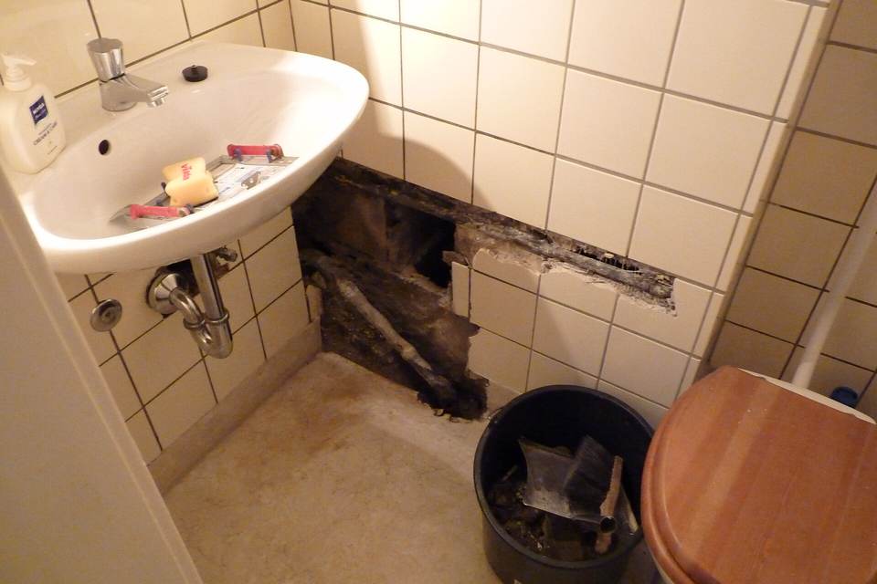 Small tiled bathroom with a strip of wall tiles removed under the sink exposing a dark wet cavity, a bucket of broken tile on the floor