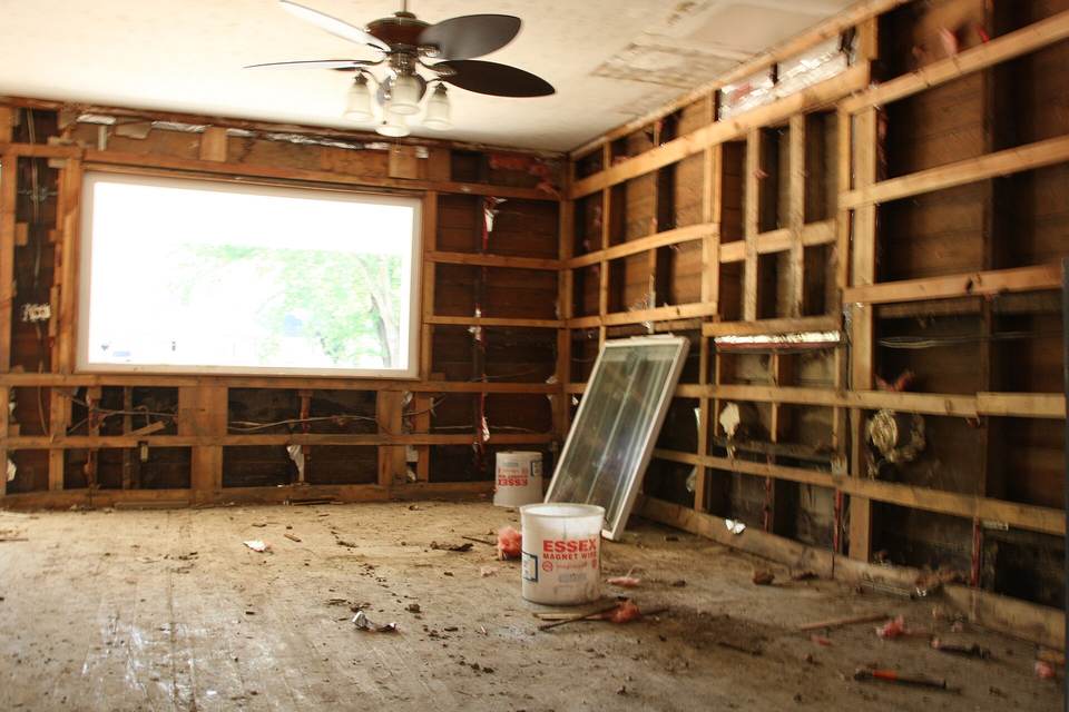 Room with every wall stripped to wood studs and wiring, a ceiling fan overhead and buckets and a window sash on a dusty floor