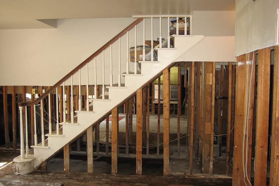 Ground floor of a house with walls stripped to bare wood studs below a white staircase that leads to the undamaged upper floor